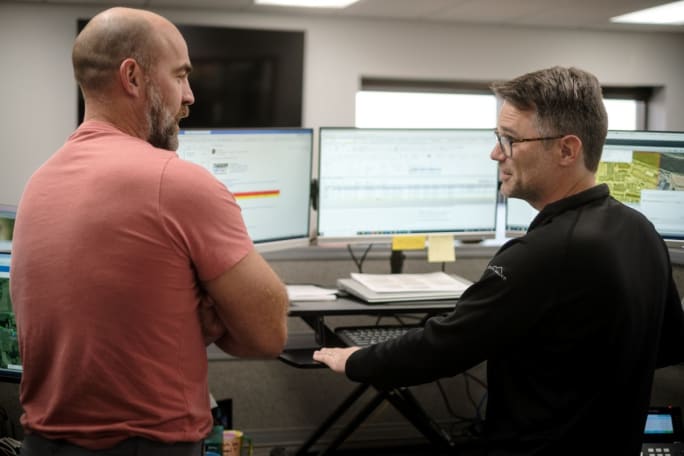 Two men in front of computer screens talking