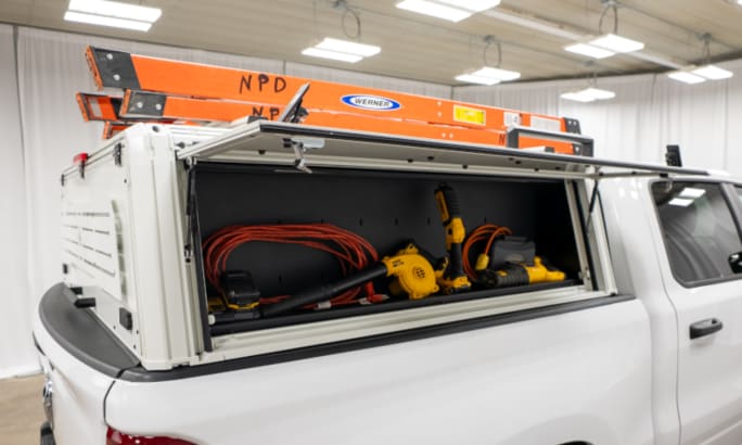 White truck cap cover installed on a white pickup truck and shown with ladders on top and the side open to reveal tool storage.