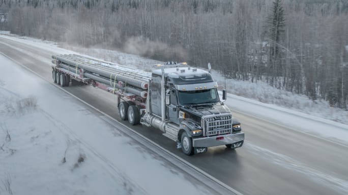Aerial view of a semi truck hauling pipes on an icy road in winter, demonstrating cold-weather endurance and durability testing in real-world conditions.