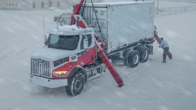 Truck with mounted crane unloading a container in heavy snow, showing equipment performance and reliability during extreme winter operations.