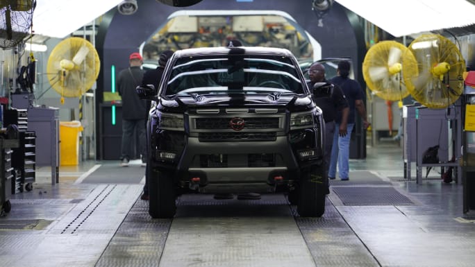Front view of a black Nissan Frontier pickup truck on the assembly line at Nissan's Canton manufacturing facility with employees working nearby.