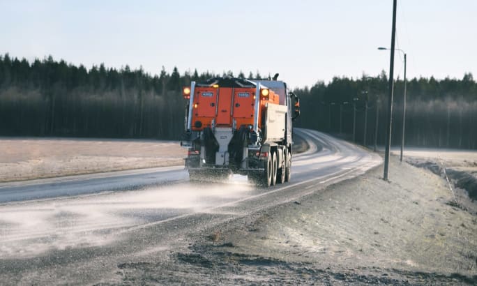 A road maintenance truck applies de-icing material to a two-lane rural roadway, spraying the pavement as it travels through a lightly frosted landscape bordered by fields and trees.