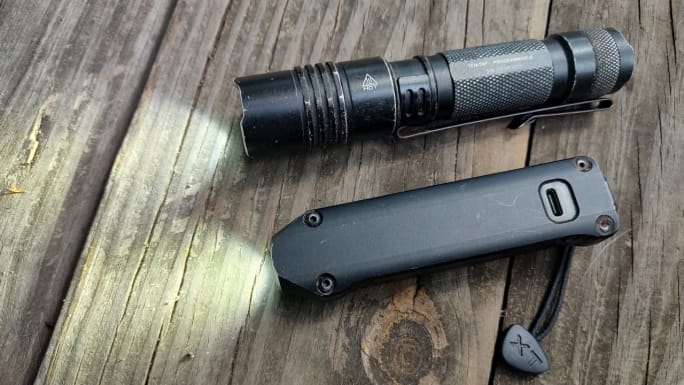 A Streamlight ProTac tactical flashlight and a Streamlight Wedge XT rechargeable EDC flashlight rest on a weathered wooden surface, comparing the traditional cylindrical design with the Wedge XT’s slim, flat profile.