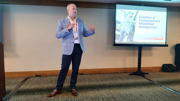 Presenter delivers a WEX conference session on connected and mixed fleet management, speaking beside a presentation screen displaying the topic, “Evolution of Connected and Mixed Fleet Management.”