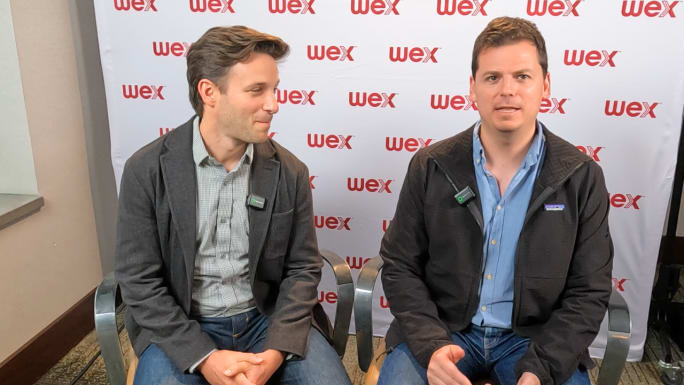 Two business professionals wearing wireless microphones sit before a WEX backdrop during an interview, with one speaking while the other listens. Both are dressed in business casual attire and seated side by side.