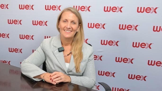 A woman wearing a wireless microphone smiles while seated at a table in front of a WEX-branded backdrop during an on-camera interview at an industry conference.