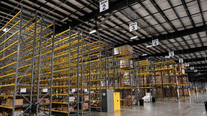 Interior of a large BendPak warehouse with tall pallet racking, labeled storage aisles, and boxed inventory organized on multiple levels beneath a high industrial ceiling, highlighting extensive parts storage and distribution capabilities.