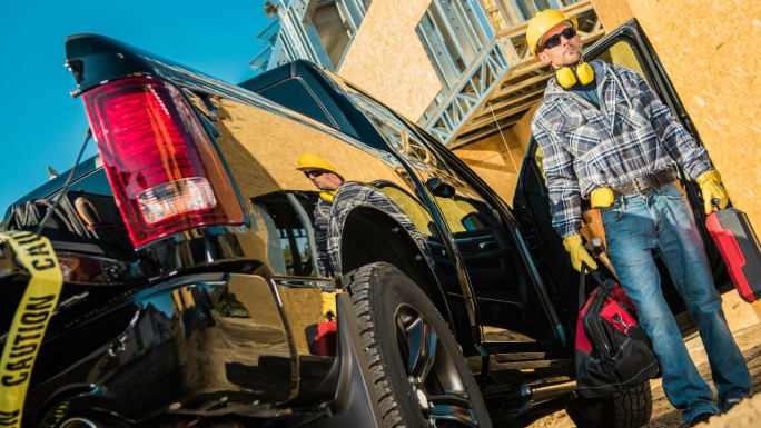 Construction worker in a hard hat stands beside an open black pickup at a jobsite, carrying tool cases and a work bag.