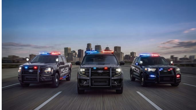 Three Ford police vehicles—a pickup, SUV, and utility vehicle—drive side by side with red and blue emergency lights flashing, with a city skyline in the background.