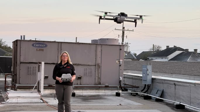A woman operates a tethered HAWK drone from a rooftop as the aircraft hovers above HVAC equipment and utility lines.