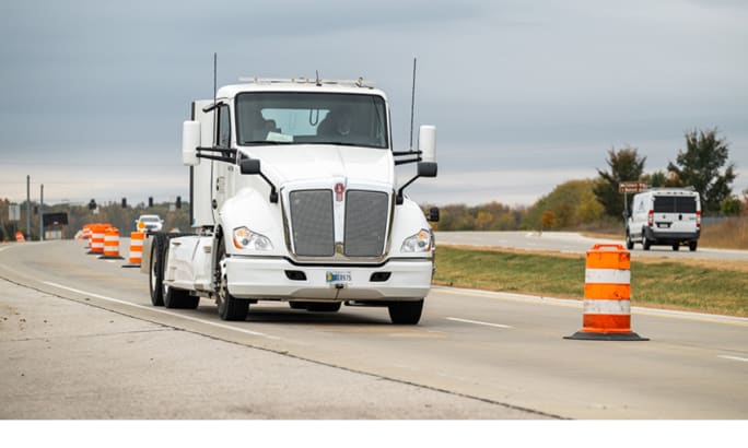 White Class 8 electric truck driving through a coned-off highway lane during wireless charging testing.