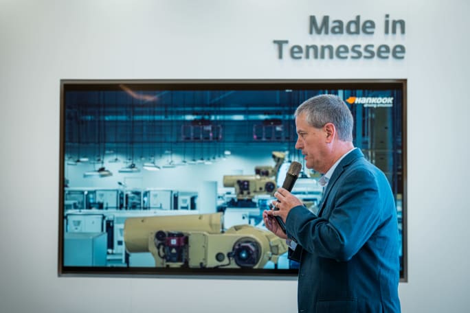 A man in a suit speaks into a microphone at a Hankook event, standing before a display showing tire manufacturing equipment beneath the words “Made in Tennessee.”