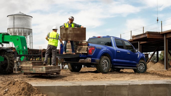 Blue Ford F-150 pickup parked at a construction site while two workers load equipment into the bed.