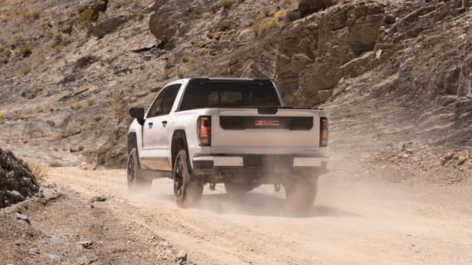 Rear view of a Coastal Dune 2027 GMC Sierra 1500 AT4 driving along a dusty off-road trail through rocky terrain.