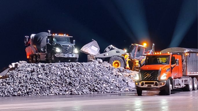 Volvo VNS vocational trucks operating at a nighttime construction site, including a concrete mixer, wheel loader, and orange tractor-trailer beside a large pile of rocks, illuminated by vehicle headlights and work lights.