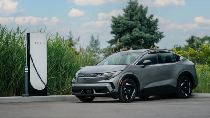 Gray 2027 Mitsubishi Eclipse Sportback connected to a Tesla charging station, with tall grasses and trees behind it.