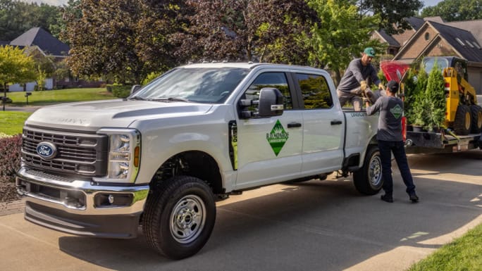 Two landscaping workers load a white Ford Super Duty Crew Cab beside a trailer carrying shrubs and a compact loader on a residential street.