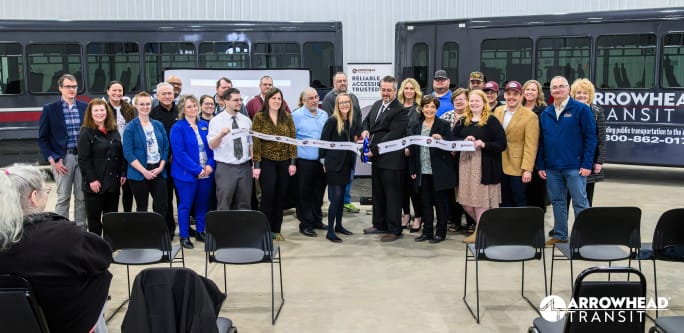 A group of people stand in front of two transit buses while a man in a suit cuts a ribbon with scissors.