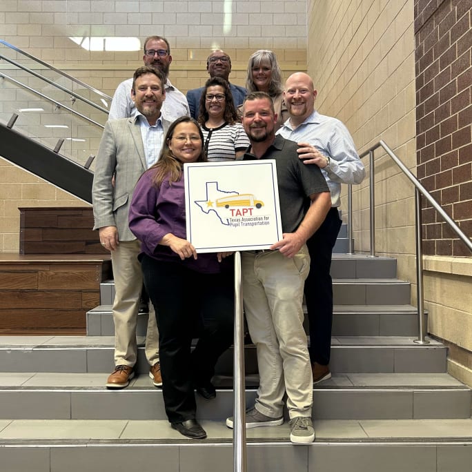 Eight people stand in a stairwell holding a Texas Association for Pupil Transportation sign