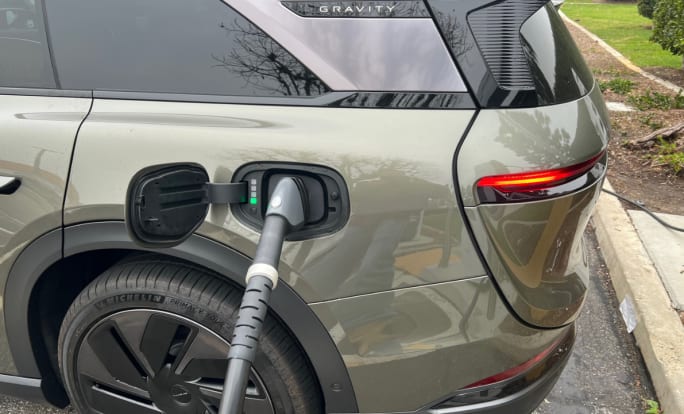 A green-gray Lucid Gravity SUV being charged at left rear charging port at a parking lot charging station.