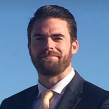 Professional headshot of a man with dark hair and a beard wearing a dark suit and tie against a blue sky.