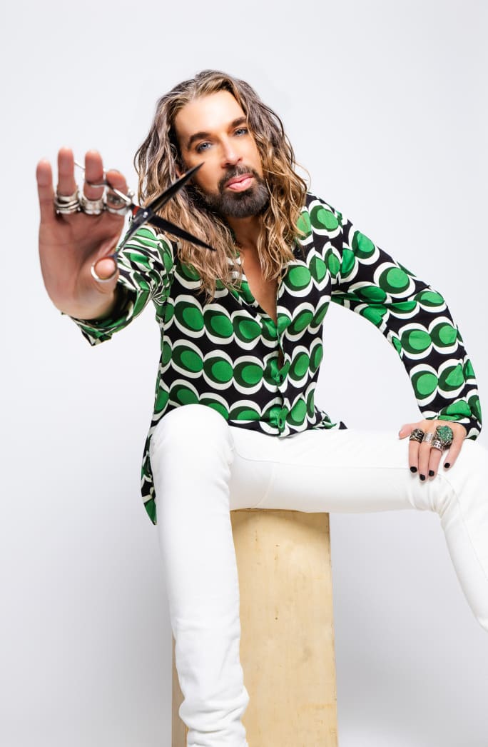 A man with long blonde hair, wearing a green shirt and white pants, holds a pair of shears toward the camera.