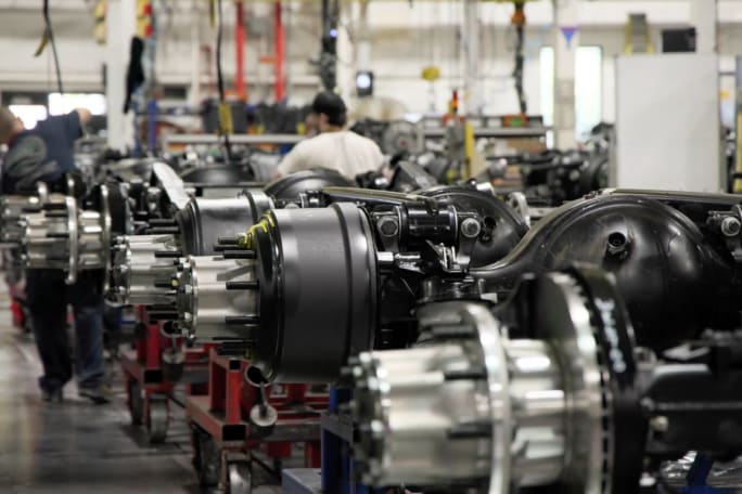 Rows of heavy-duty truck axle assemblies on carts inside a Daimler Truck North America manufacturing plant, with workers assembling components in the background.