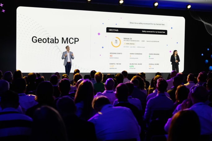 A man and a woman on stage in a darkened convention room, backed by a large screen illustrating Geotab MCP