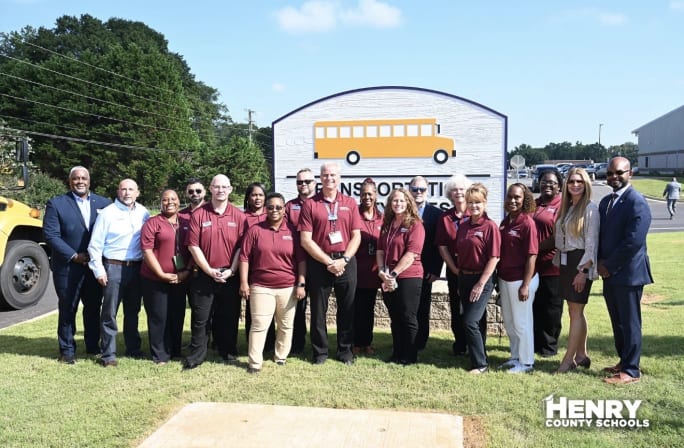 A group of transportation employees stand in front of a Henry County Schools sign outside on a sunny day