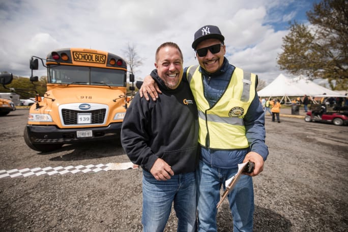 Two men smile with their arms around each other in front of a yellow school bus at an outdoor New York school bus competition event. One wears a reflective safety vest and holds a clipboard.