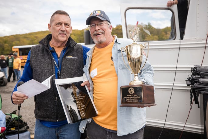 Two men smile while posing with awards at the New York State School Bus Safety Competition, with one holding a first-place trophy and a Rookie of the Year plaque.