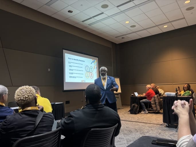 Bernando Brown presents to a group session at an NAPT conference with a slideshow on a projector screen behind him.