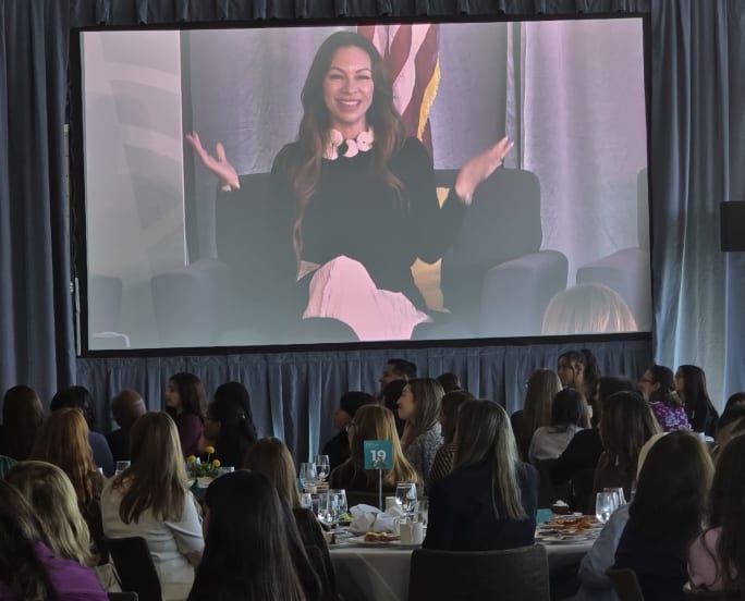 Jennie Abarca on a large video screen in front of a group of people sitting at tables