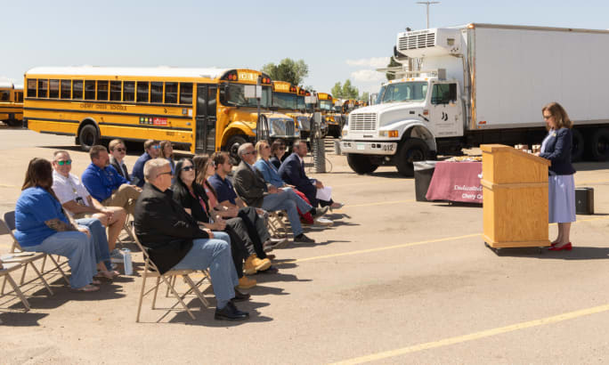 A small crowd of people sitting during a speech at an outdoor electric school bus depot groundbreaking.