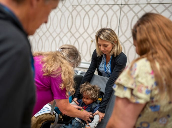 Katia Dubas of IMMI demonstrates a child safety restraint with a toddler seated in a bus seat while transportation professionals observe during a hands-on school bus safety training.