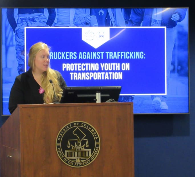 Woman speaking at a podium with the District of Columbia Office of the Attorney General seal, presenting “Truckers Against Trafficking: Protecting Youth on Transportation” on a large screen behind her.