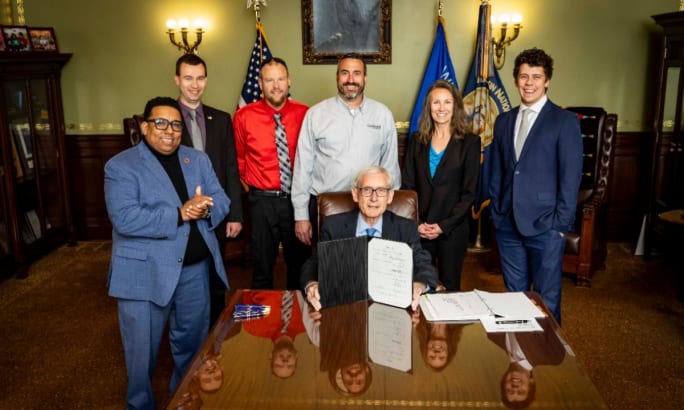 Wisconsin Governor Tony Evers sits at a table with people standing behind him.