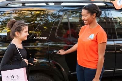  a woman in an orange shift opens the door for a student next to a black minivan