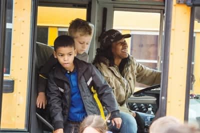  Two students exit a yellow school bus as the smiling bus driver sits behind the wheel.