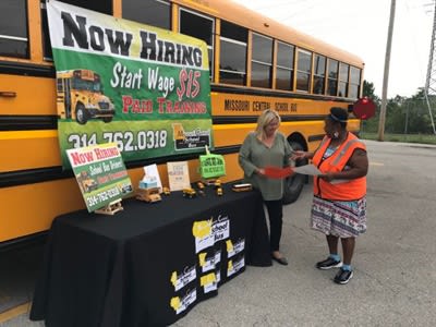  Two women stand at a school bus driver recruitment table beside a yellow school bus displaying a “Now Hiring” banner with starting wage and paid training information.