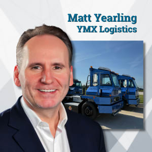 Man in suit jacket with blue electric yard tractors in background