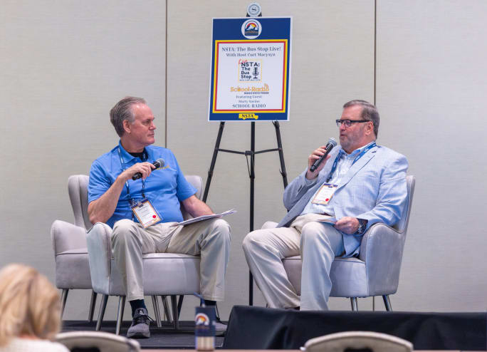 NSTA Executive Director Curt Macysyn interviews Marty Savino of School Radio during a live recording of NSTA: The Bus Stop podcast at the 2026 NSTA Annual Meeting & Convention in Denver. The two sit onstage with microphones as attendees watch from the audience.