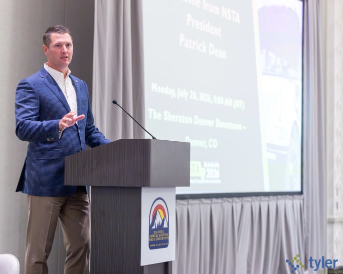 NSTA President Patrick Dean delivers opening remarks from the podium during the 2026 NSTA Annual Meeting & Convention in Denver. A presentation screen behind him displays the conference agenda as he welcomes attendees to the event.