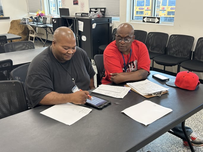 Two men sit at a table with papers in front of them.