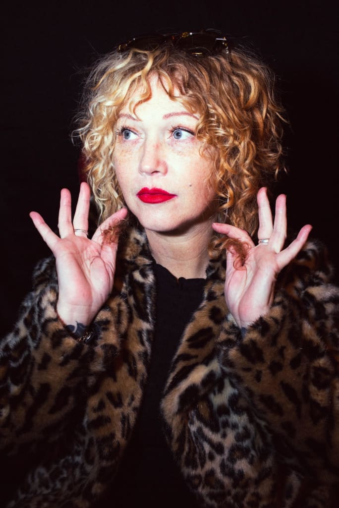 A woman with red curly hair, wearing a leapoard print coat, holds pieces of her hair out.