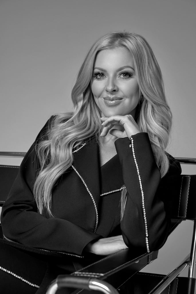 A woman with long blonde hair, posing with her hand under her chin.