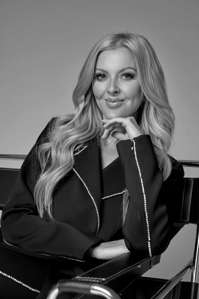 A woman with long blonde hair, posing with her hand under her chin.
