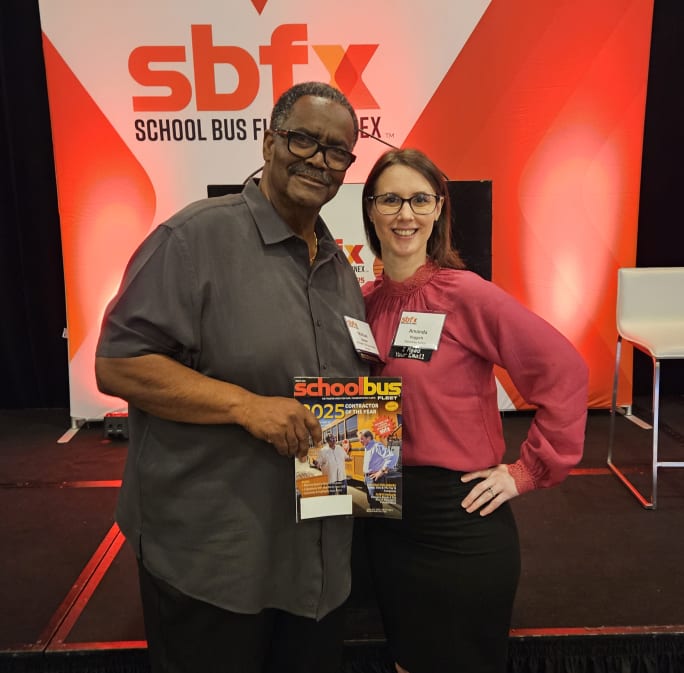 Two people pose at the School Bus Fleet Exchange stage, holding a School Bus Fleet magazine, with SBFX branding and red stage lighting in the background.