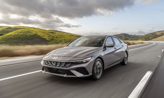 A gold/gray 2025 Hyundai Elantra driving down a paved road with mountains in the background.