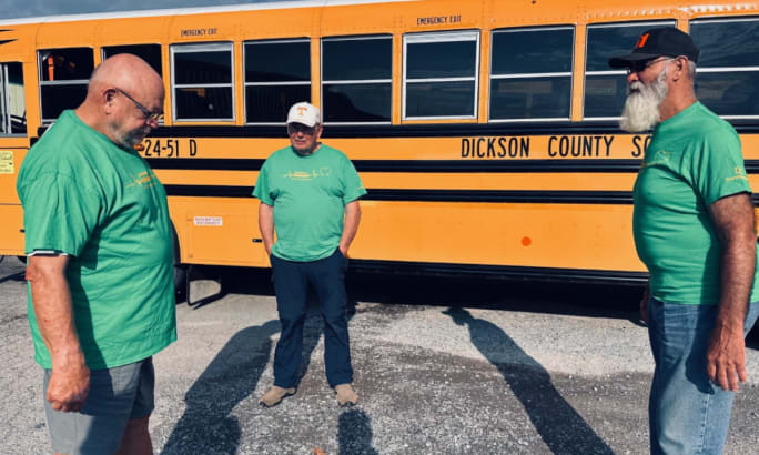 Three men in green shirts talk next to a school bus.